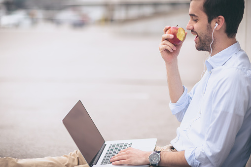 Young man is eating an apple  while on the brake. He is wearing white shirt and khaki pants. He is wearing beard and slicked hair. The photography taken with shallow depth of field, focus on apple