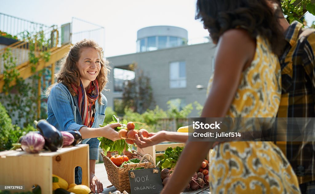 Friendly woman tending an organic vegetable stall at a farmer Female gardener selling organic crops and picking up a bountiful basket full of fresh produce Agricultural Fair Stock Photo