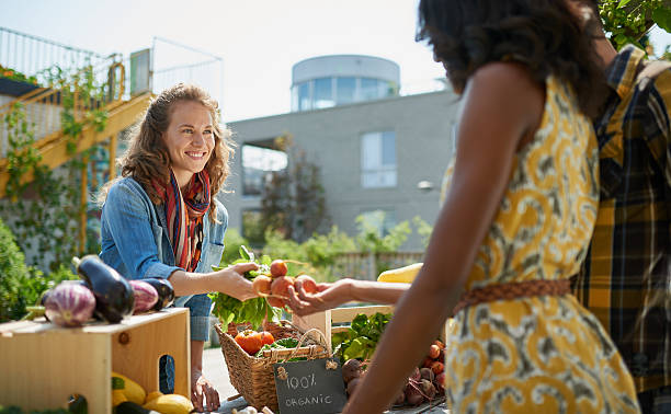 Friendly woman tending an organic vegetable stall at a farmer Female gardener selling organic crops and picking up a bountiful basket full of fresh produce farmers market stock pictures, royalty-free photos & images