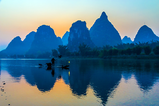 Silhouette of Man fishing with Cormorant Bird floating on traditional Boat in Central China River unusual shaped rocky Mountains and Morning Sky on Background