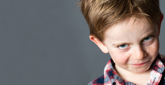 closeup portrait of a young mischievous child with freckles teasing and grumbling with fun look and joke, copy space on grey background studio