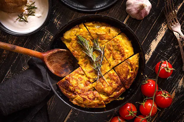 Tortilla de patatas in a pan with garlic sauce aioli and fresh tomatoes cherry. Traditional spanish dish. Omelette with eggs, potatoes and onion. Rustic black background. Top view