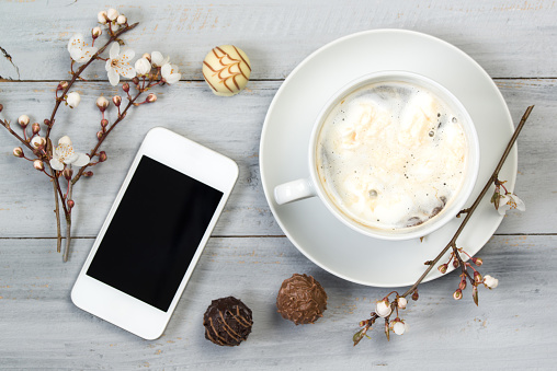 Cup of coffee with cream and smartphone, on wooden table with cherry flowers and chocolate candy, top view