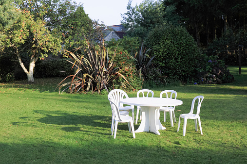 White plastic garden furniture catches the evening sun in a garden in Brittany, France. Long shadows are on the lawn. Fruit trees and other plants provide a backdrop