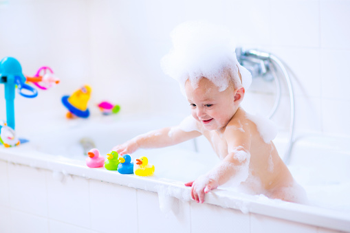 Cute little baby boy taking bath playing with foam and colorful rubber duck toys in a white sunny bathroom