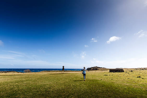 A DSLR photo taken in Easter Island, Chile. A young tourist is walking in a huge open grass area towards a single Moia statue. Is it a bright sunny day with blue sky and scattered white clouds.