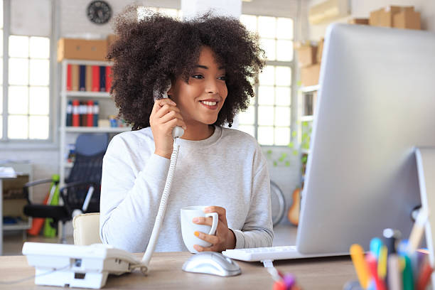 Smiling businesswoman talking on the landline phone Happy young businesswoman holding an earphone of landline phone and a cup and looking at computer.Tall windows and shelfs with boxes and folders as background. Landline phone, mouse, keyboard and tools on desk landline phone stock pictures, royalty-free photos & images