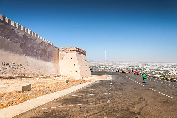 A scenic view of Rabat's historic fortified wall, surrounded by palm trees and a clear blue sky. People stroll the vibrant park area, enjoying the city's architectural and cultural landmarks.