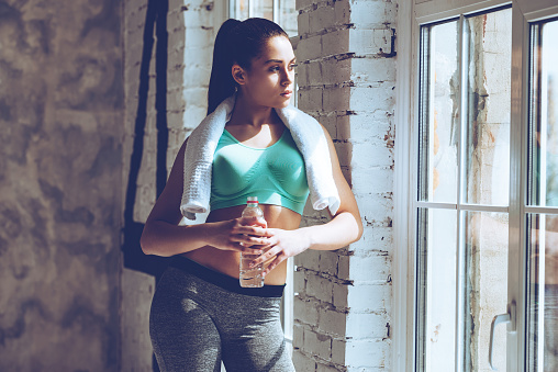 Beautiful young woman with towel on her shoulders holding water bottle and looking through window while leaning to the wall at gym