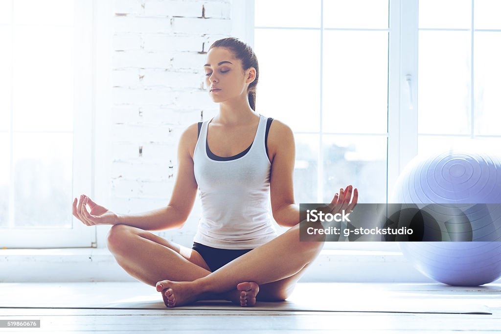 Namaste! Young beautiful young woman keeping eyes closed while sitting in lotus position in front of window at gym Meditating Stock Photo