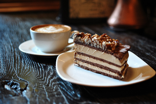 chocolate cream cake with nuts on a plate and a cup of cappuccino coffe on a black table