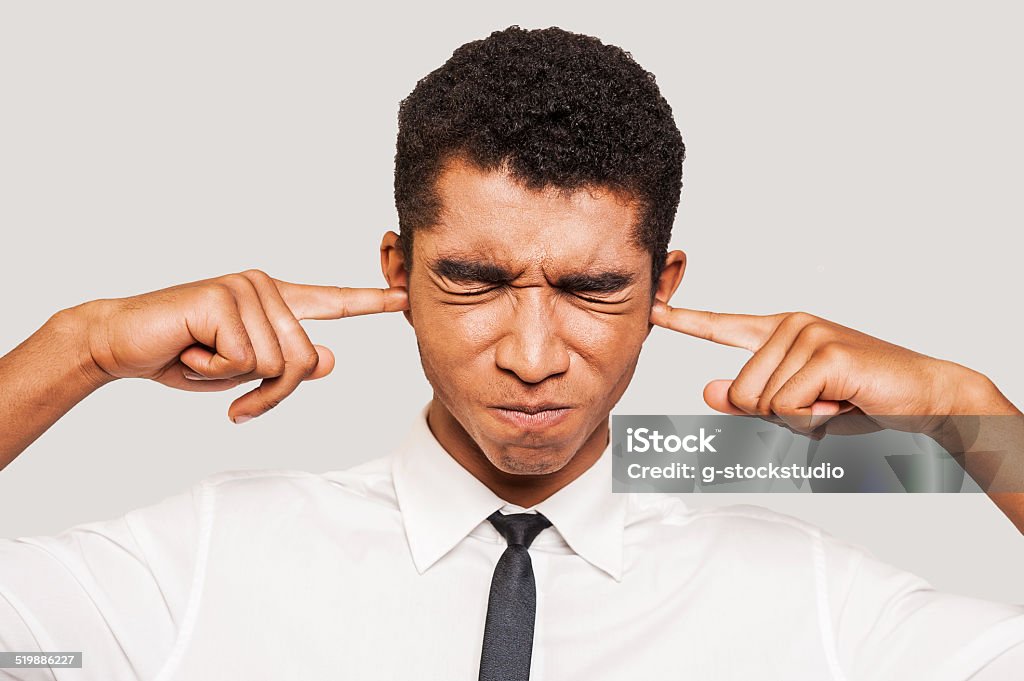 This is too loud! Frustrated young Afro-American man in formalwear plugging ears with his fingers and keeping eyes closed while standing against grey background Adult Stock Photo