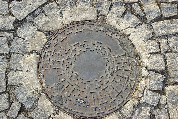 Well Lid Closeup in Cobblestone Way Cobblestone Path with Iron Cover. Background and Texture for text or image sewer lid stock pictures, royalty-free photos & images