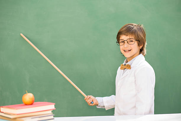 Education, children.  Happy boy in elementary classroom. Cute little boy wearing bow tie, glasses, and lab coat is all smiles as he uses pointer on a green chalkboard.  The elementary age, Latin descent child is playing dress up as a professor, is excited about the first day of school, or is proud of his recent classroom success.  Copyspace to side.  Apple, books stacked. pointer stick stock pictures, royalty-free photos & images