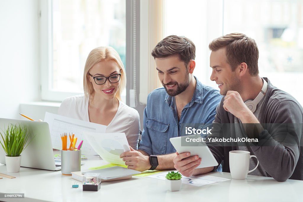 Cheerful young colleagues are doing paperwork Skillful creative team is working with documents. They are sitting at table and smiling. Man is holding a tablet Adult Stock Photo