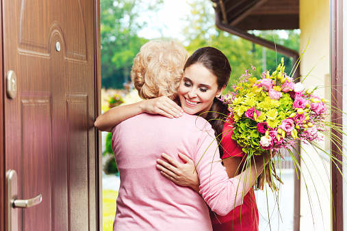 Happy young woman bringing birthday flowers to senior woman, standing at the entrance door and hugging. 