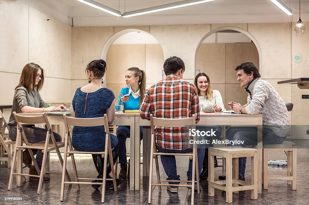 Group of Students sitting at Campus Club Area and discussing Many Students in casual Clothes Men and Women sitting at wooden Table inside Campus Chat Room area talking laughing working Entertainment Club Stock Photo