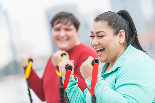 An overweight Hispanic woman in her 30s exercising outdoors with resistance bands. Her friend is out of focus in the background. She is laughing, having fun.