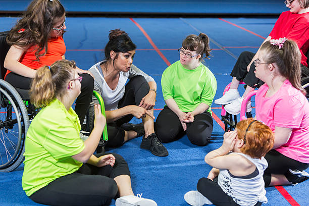 Group of special needs girls with instructor in gym An instructor with a group of special needs teenagers and young women, sitting in a circle on the floor of a gym, talking. The girls have various mental and physical disabilities, including down syndrome and morquio syndrome. Two of the teens are in wheelchairs. Disability Support Worker Course stock pictures, royalty-free photos & images
