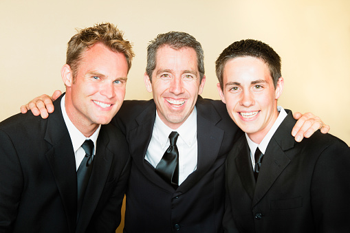Three smiling men in black suits portrait in front of beige background.