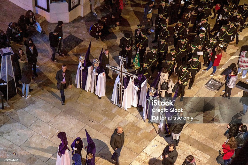 Semana Santa in Malaga: procession of Rico brotherhood Malaga, Spain - March 24, 2016: nightly return of the Easter procession of the Rico brotherhood through Calle Alcazabilla, headed by nazarenos, or penitents, carrying a large cross and other ritual objects. They are dressed in purple robes and pointed hoods with white capes. The nazarenos are followed by a brass band. Andalusia Stock Photo