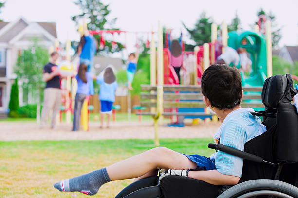 little boy para personas con discapacidades en silla de ruedas, mientras ve los niños juegan en el patio de juegos - ostracized fotografías e imágenes de stock