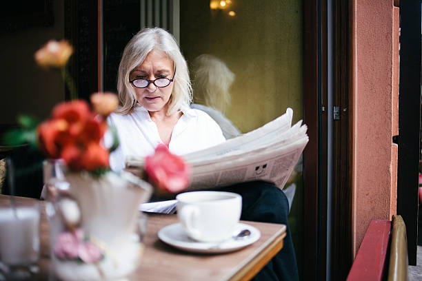 senior mujer está ubicado en la terraza de un café y lee el periódico - mujer leyendo periodico fotografías e imágenes de stock