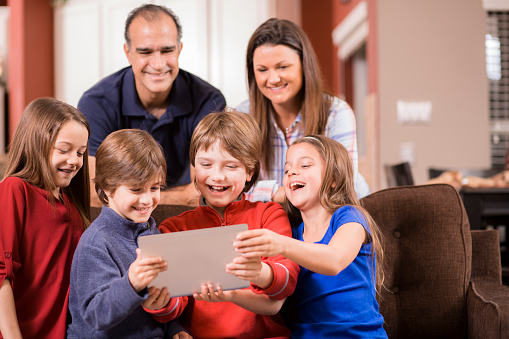 Mixed race family of six using a digital tablet at home.  The Latin and Caucasian family members are sitting on the living room sofa as they enjoy a movie, video or video chat together.  Kitchen in background.  Mother, father, two girls, and two boys children. Happy, laughing, smiling.