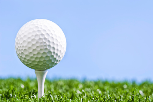 A low angle view of a Golf ball sitting on white wooden tee against a blue sky, with grass underneath 