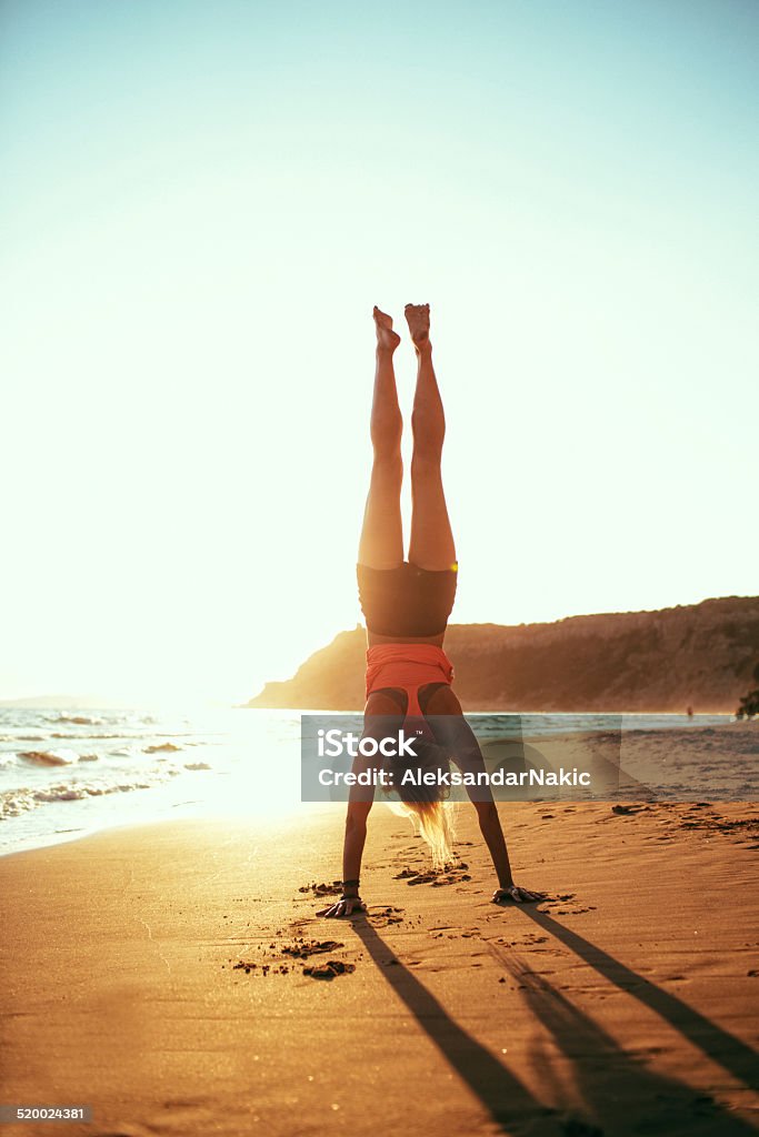 Young woman practising yoga on the beach Young woman practising yoga on the beach at sunset, performing handstand pose Adult Stock Photo