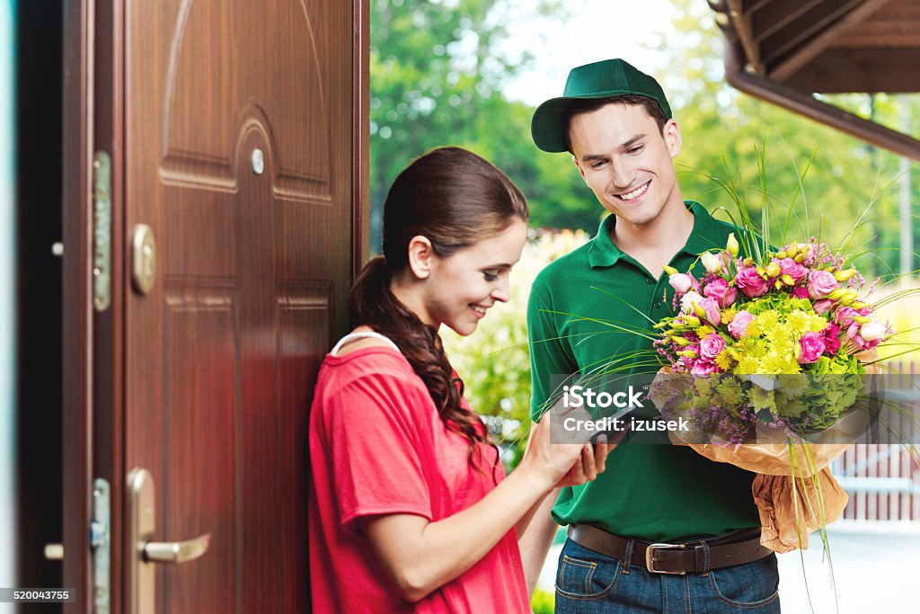 Young delivery man delivering flowers Smiling delivery man delivering a bouquet of flowers for young woman, standing in th entrance house door and woman signing up on the palmtop.  Delivering Stock Photo