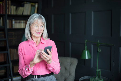 Portrait of senior woman with mobile phone in living room. Woman at home texting on cell phone, smiling and looking away.