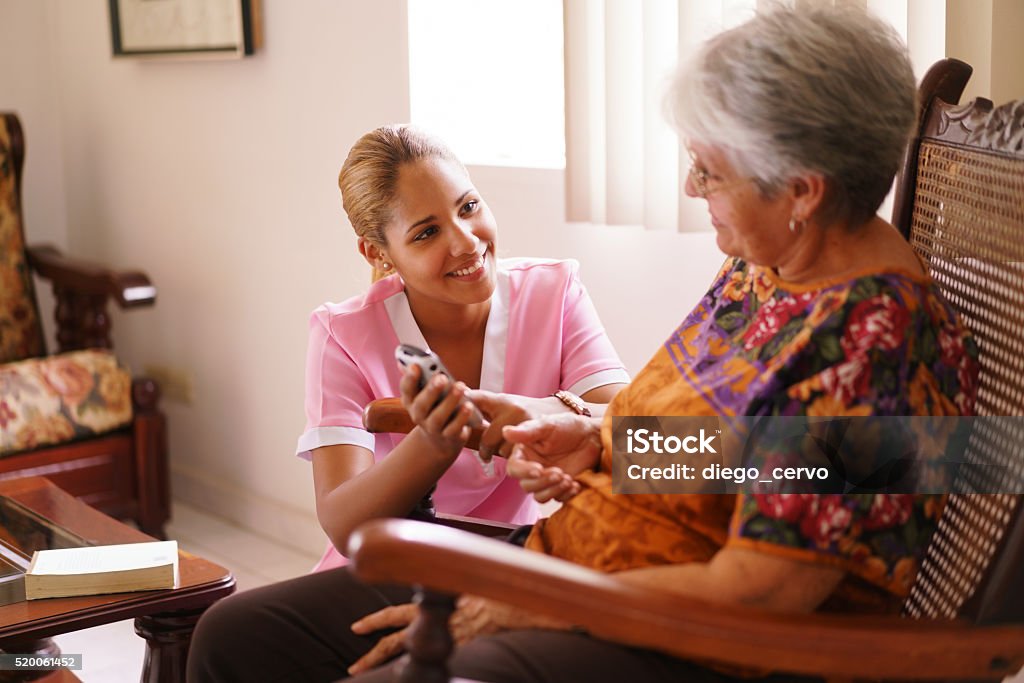 Hospice Nurse Helps Old Lady With Mobile Phone Call Old people in geriatric hospice: elderly lady having eyesight problems viewing the screen of mobile phone. A nurse helps the senior woman dialing a number on the tiny keyboard Latin American and Hispanic Ethnicity Stock Photo