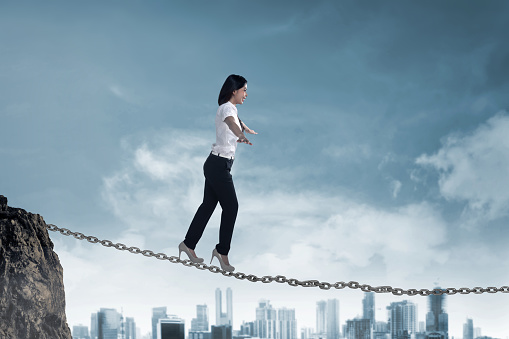 Asian business person balancing on old iron chain with cloudy sky background