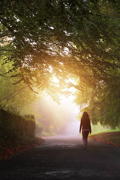 A woman walks towards the light on a misty day. A solitary woman walking on a country road