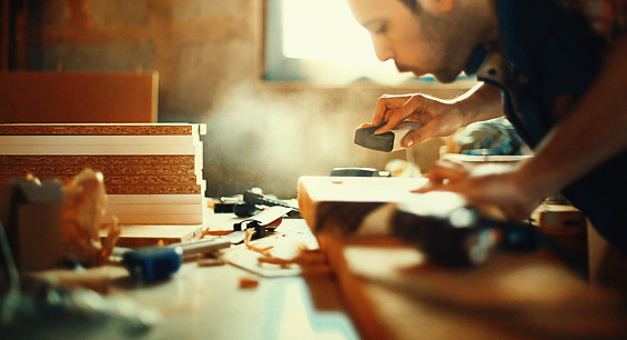Closeup of mid 30's carpenter sanding a piece of wood. He's blowing sawdust after few strokes. Back lit, oned image.