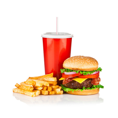 Cheeseburger, french fries and cola drink isolated on reflective white backdrop, typical combination for this meal. A red disposable cup of cola is in the center, the fries are laying at the left of the cola and a huge cheeseburger is at the right. The ingredients of the burger are a big beef portion, lettuce, bacon, tomato, and bacon on sesame seed bun.