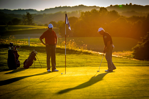 Golf pro teaching male golfer on putting green at sunset.