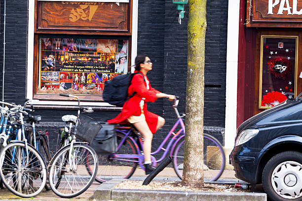 Amsterdam, The Netherlands - June 9, 2014: An Asian woman dressed in red coat is cycling in a rush along canal Vorburgwal in Amsterdam in summer. She is carrying a huge backpack. At canal are some bicycles and a car. In background are restaurants and bars.