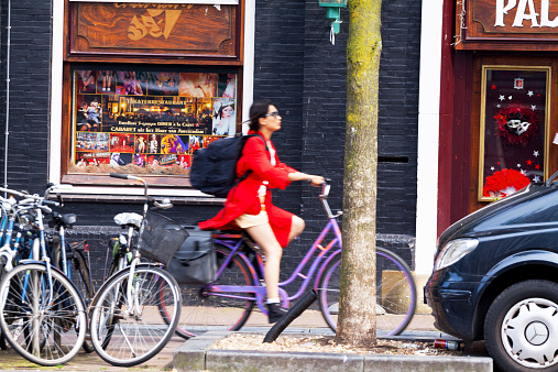 Amsterdam, The Netherlands - June 9, 2014: An Asian woman dressed in red coat is cycling in a rush along canal Vorburgwal in Amsterdam in summer. She is carrying a huge backpack. At canal are some bicycles and a car. In background are restaurants and bars.