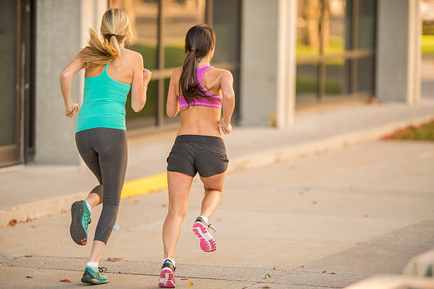 Girlfriends Running Together Two girlfriends are wearing athletic clothing and is working out by going on a jog through the city. spandex stock pictures, royalty-free photos & images