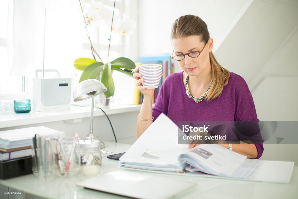 Woman working at home. Woman working in her attic home office, with big window and natural light. Sitting at the table, wearing glasses and elegant clothes. Looking through some documents while holding cup go coffee. Office colour is grayish white. Nordic style. Copenhagen, Denmark.  Reading Stock Photo