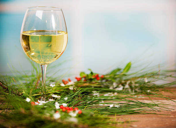 White Christmas wine glass on top of pine tree needles, holly, and snowflakes. Wooden table and blue window background. Rustic.