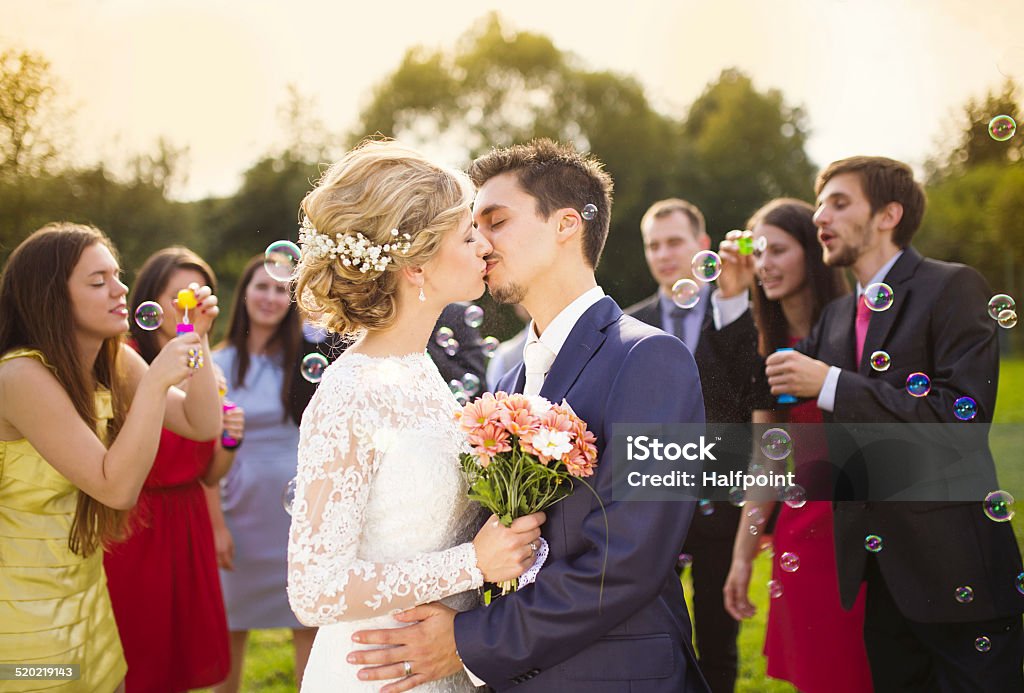 Newlyweds kissing at wedding reception Young newlyweds kissing and enjoying romantic moment together at wedding reception outside, wedding guests in background blowing bubbles Wedding Ceremony Stock Photo