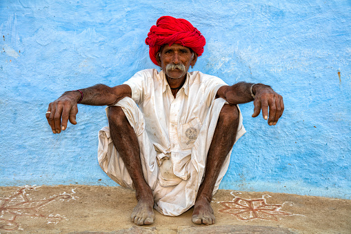 Khairwa, India - December 4, 2015: Senior Rajasthani man with red turban on the head sitting in front of blue painted wall of his house in Khairwa, Rajasthan, India. Khairwa is a village in Pali district.