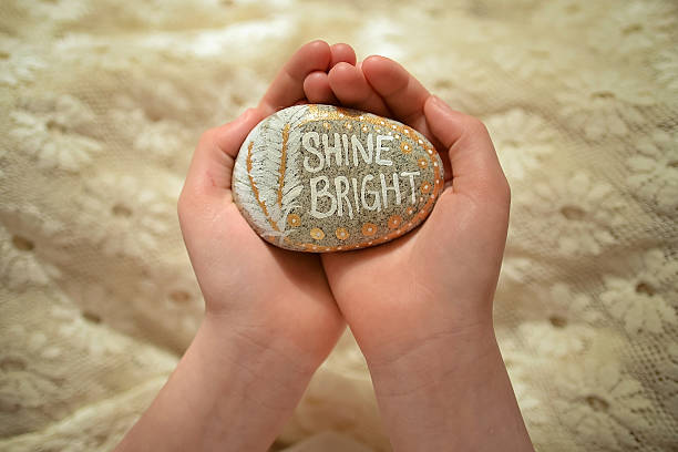 Two hands hold a rock with the words Shine Bright painted onto it.  These are uplifting, therapy rocks.  Shine Bright is painted in white, there are two painted feathers and gold details.  The background is a white lace fabric with flowers.