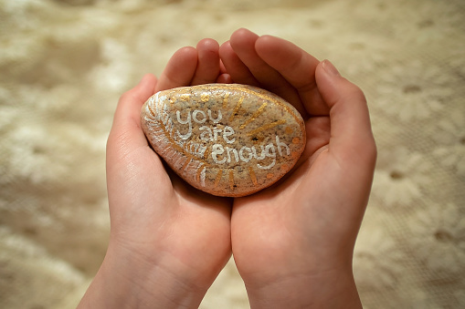 Hands hold a beautiful painted rock with the words You Are Enough painted on it.  The hands are offering us this sentiment.  The words are painted in white with a hand painted feather and gold paint details
