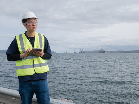 An asian engineer holding a tablet and standing in front of semi submersible oil rig