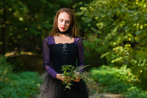 young woman in purple blouse, black full length dress and black corset holding wild flowers, looking downwards