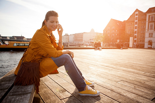 Woman wearing orange coat sitting on bench near canal of Copenhagen and talking on smart phone. Sunset, evening. Wooden floor, buildings in background. Nyhavn , Copenhagen, Denmark.